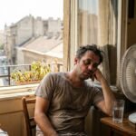 Hőségben fáradt ember árnyékos szobában, vízzel és ventilátorral próbálja lehűteni magát